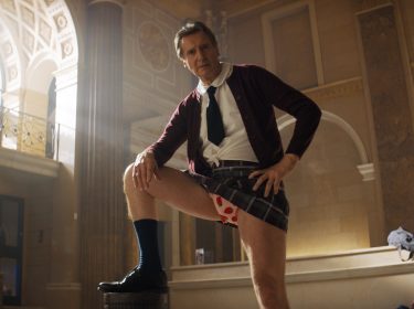 In dieser Szene aus Die nackte Kanone 4 von 2025 steht Liam Neeson als Frank in einem prachtvollen, klassizistischen Gebäude mit einem Bein auf einem Podest. Er trägt eine Schuluniform mit offenem Hemd, Krawatte, kariertem Rock und sichtbar roten Herzchen-Boxershorts, was der Szene eine absurde und humorvolle Stimmung verleiht.