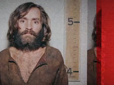 Charles Manson wird im Gefängnis frontal fotografiert, während er einen braunen Longsleeve trägt.