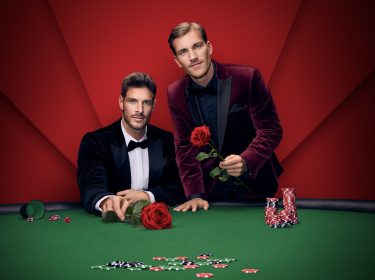 Auf dem Bild zu Die Bachelors 2025 stehen zwei stilvoll gekleidete Männer an einem Pokertisch mit grüner Filzfläche. Beide tragen elegante Samt-Sakkos, der linke in Dunkelblau mit Fliege, der rechte in Weinrot. Beide halten jeweils eine rote Rose in der Hand. Auf dem Tisch liegen Pokerchips und Würfel, der Hintergrund ist rot inszeniert.