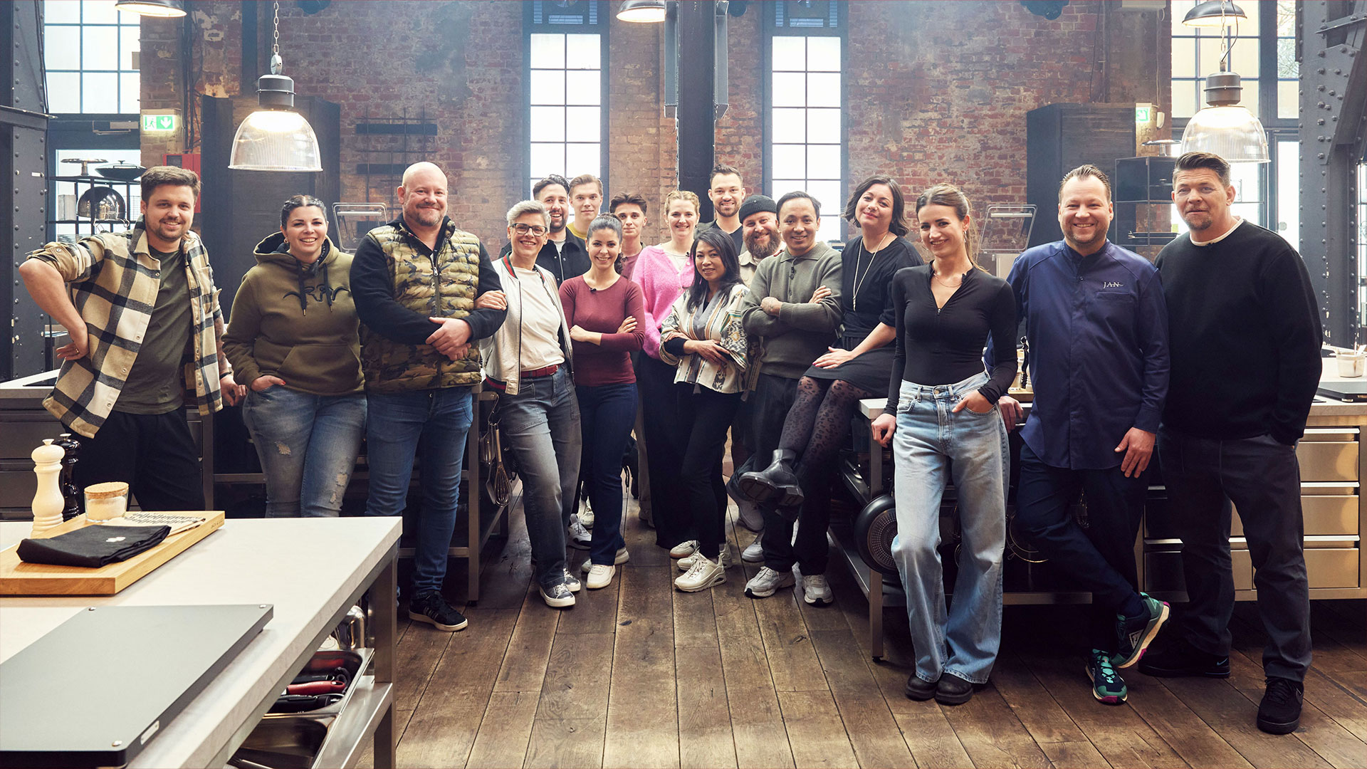 Auf dem Bild zur Sendung Mälzers Meisterklasse VOX steht eine Gruppe von Teilnehmer:innen gemeinsam mit Tim Mälzer in einer modernen Studioküche. 