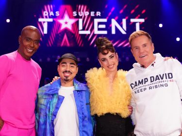 Auf dem Bild zur Show Das Supertalent 2025 ist die Jury mit Bruce Darnell, Tony Bauer, Ekaterina Leonova und Dieter Bohlen zu sehen. Vor einer leuchtenden Bühne mit dem Show-Logo posieren sie lächelnd für die Kamera. Bruce Darnell trägt ein auffälliges pinkes Outfit, Tony Bauer eine blaue Jacke, Ekaterina Leonova ein gelbes Tüll-Oberteil und Dieter Bohlen einen weißen Hoodie mit 