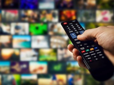 Eine Hand hält eine schwarze Fernbedienung und navigiert durch eine Auswahl an Streamingdiensten in Deutschland. Im Hintergrund sind zahlreiche unscharfe Bildschirmvorschauen von Filmen und Serien zu sehen, die eine große Vielfalt an Inhalten darstellen. Die Szene vermittelt das moderne Fernseherlebnis mit On-Demand-Streaming.