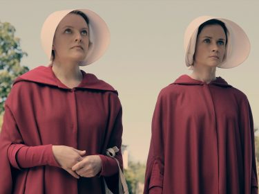 Auf dem Bild in dem Artikel zu The Testaments sind zwei Frauen in roten Umhängen und weißen Hauben zu sehen, die ernst und entschlossen in die Ferne blicken. Die Szene spielt in einer natürlichen Umgebung mit Bäumen im Hintergrund und vermittelt eine bedrückende, aber kämpferische Stimmung.