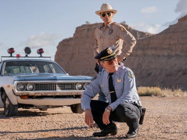 In Dark Winds Staffel 3 sind Lt. Joe Leaphorn (Zahn McClarnon) und Gordo Sena (A. Martinez) in einer wüstenartigen Landschaft zu sehen. Leaphorn steht mit Sonnenbrille und Hut neben einem alten Polizeiwagen, während Sena in blauer Uniform und mit ernster Miene in der Hocke verharrt.