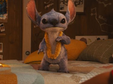 Auf dem Bild aus der Lilo und Stitch Live-Action-Adaption 2025 ist das blaue Alien Stitch in einer realistischen CGI-Version zu sehen. Er steht auf einem Bett, trägt eine gelbe Blumenlei und blickt direkt in die Kamera.