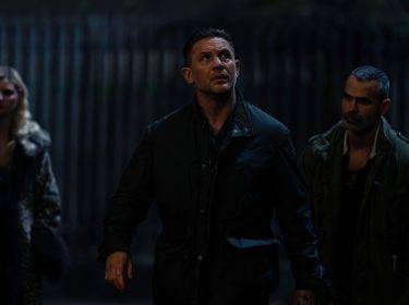 Auf dem Bild zur Mobland Serie ist Tom Hardy als Harry Da Souza zu sehen. Er steht in einer dunklen Gasse und blickt angespannt nach oben, als ob er eine Bedrohung wahrnimmt. Er trägt eine dunkle Jacke, und sein Gesichtsausdruck wirkt fokussiert. Hinter ihm stehen zwei weitere Personen – eine Frau mit blondem Haar in einem auffälligen Mantel und ein Mann mit kurz rasiertem Haar und einem ernsten Blick. Die Szene ist düster beleuchtet und vermittelt eine gespannte Atmosphäre.