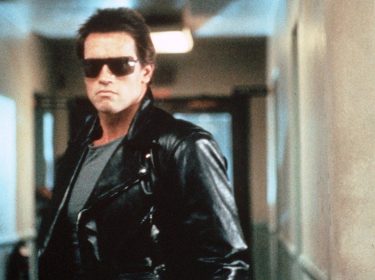 Auf dem Bild aus Terminator ist Arnold Schwarzenegger in seiner ikonischen Rolle als der Terminator zu sehen. Er trägt eine schwarze Lederjacke, eine dunkle Sonnenbrille und ein graues Shirt, während er mit ernstem Blick durch einen Flur geht. Die düstere Beleuchtung und sein ausdrucksloses Gesicht verstärken die bedrohliche Atmosphäre der Szene.