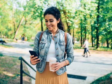 Frau steht im Park mit ihrem Smartphone und einem Kaffee in der Hand.