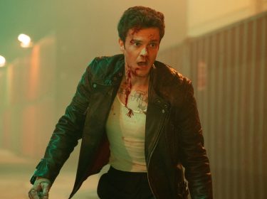 Auf dem Bild aus der Mr. No Pain Kritik ist Jack Quaid als Nathan Cain zu sehen. Er trägt eine schwarze Lederjacke über einem blutbefleckten weißen Unterhemd und wirkt erschöpft sowie alarmiert. Sein Gesicht ist mit Blutspuren bedeckt, und er scheint sich in einem beleuchteten Containerlager zu befinden. Die düstere Atmosphäre und sein angespannter Ausdruck deuten auf eine dramatische Szene voller Action hin.