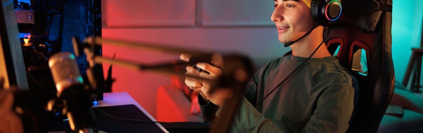 Ein junger Mann sitzt auf einem PC-Stuhl, hält einen Controller in der Hand und spielt am Computer.