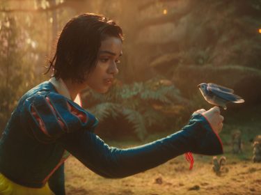 Auf dem Bild in dem Artikel zu der Schneewittchen-Filmkritik ist Schneewittchen, gespielt von Rachel Zegler, in einem märchenhaften Wald zu sehen. Sie trägt ihr ikonisches blau-gelbes Kleid und hält vorsichtig die Hand ausgestreckt, auf der ein blauer Vogel sitzt. Ihr Blick ist sanft und nachdenklich, während im Hintergrund kleine Waldtiere zu erkennen sind. Die Szene ist in warmes, goldenes Licht getaucht, was eine magische Atmosphäre erzeugt.