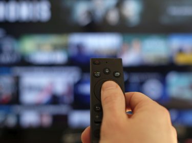 Hand hält eine Fernbedienung vor einen Fernseher