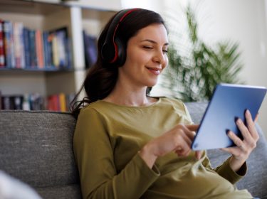 Auf dem Bild ist eine junge Frau mit dunklen Haaren zu sehen, die entspannt auf einem grauen Sofa sitzt. Sie trägt schwarze Kopfhörer mit roten Akzenten und ein olivgrünes Oberteil. In ihren Händen hält sie ein blaues Tablet und tippt darauf, während sie zufrieden lächelt. Im Hintergrund steht ein Bücherregal mit bunten Büchern. Das Bild vermittelt eine gemütliche Atmosphäre, passend zur Nutzung von Arthaus Plus für Streaming.