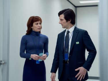 Auf dem Bild aus Severance Staffel 2 sind Adam Scott als Mark Scout und Britt Lower als Helly Riggs in einem sterilen, weiß beleuchteten Büroflur zu sehen. Mark trägt einen dunklen Anzug mit Krawatte, während Helly ein eng anliegendes blaues Rollkragenkleid trägt. Beide tragen ID-Karten an blauen Bändern und führen ein ernstes Gespräch. Severance Staffel 2 Ende erklärt – diese Szene deutet auf eine wichtige Entwicklung innerhalb von Lumon Industries hin.