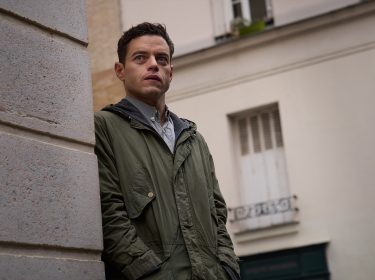 Auf dem Bild aus The Amateur ist Charles Heller zu sehen, gespielt von Rami Malek. Er steht in einer Straßenszene an einer Häuserwand und blickt mit ernstem, nachdenklichem Gesichtsausdruck in die Ferne. Sein olivgrüner Parka und die gedeckten Farben der Umgebung verleihen der Szene eine gespannte, geheimnisvolle Stimmung. Das Bild vermittelt einen stillen Moment voller innerer Anspannung und Ungewissheit.