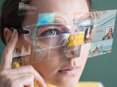 Auf dem Bild zum Thema Was ist Augmented Reality ist eine Frau zu sehen, die eine futuristische AR-Brille trägt. Vor ihrem Gesicht schweben holografische Benutzeroberflächen mit Begriffen wie „Search“, „Business“, „Technology“ und „Vacation“. Die Darstellung vermittelt eine technologische, zukunftsorientierte Stimmung und zeigt, wie Augmented Reality digitale Inhalte in die reale Umgebung integriert.