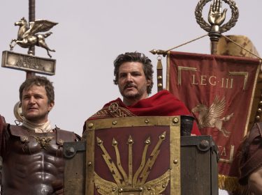 Auf dem Bild aus Gladiator 2 ist Pedro Pascal als Marcus Acacius in einer eindrucksvollen Szene in römischer Rüstung zu sehen. Er steht mittig mit ernstem Blick und rotem Umhang vor dem Banner der dritten Legion.