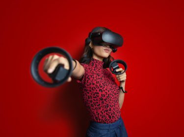 Eine junge Frau mit Virtual-Reality-Brille und zwei Controllern streckt den rechten Arm energisch nach vorne. Sie trägt ein rotes Oberteil mit Leopardenmuster vor knallrotem Hintergrund. Der Ausdruck wirkt fokussiert und spielerisch. Das Bild veranschaulicht auf moderne Weise die zentrale Frage: Was ist Virtual Reality?