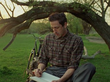 GigaTV-Highlights im April 2025: In einer Szene aus The Brutalist sitzt Adrien Brody als zurückhaltender Architekt in einem ruhigen Park auf dem Gras und schreibt in ein Notizbuch.