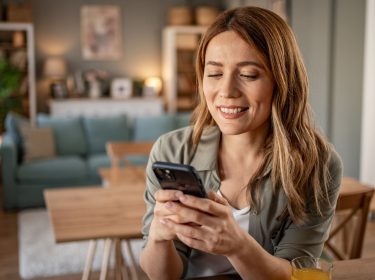 Eine Frau sitzt in einer Wohnung an einem Tisch, hält ein Smartphone in der Hand und lächelt.