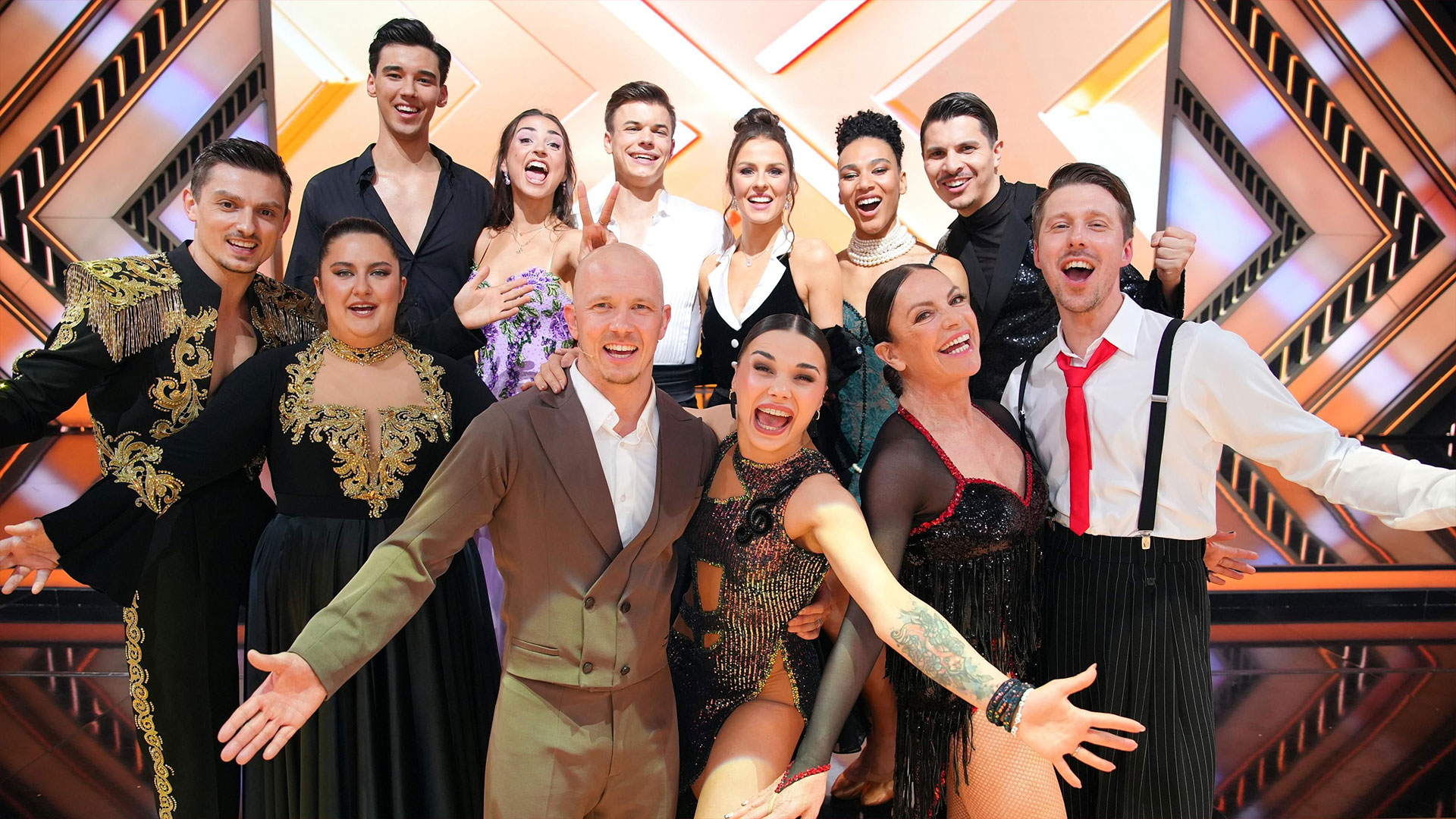Auf dem Bild zur Let's Dance 2025 Show 9 sind die Tanzpaare der RTL-Show zu sehen. Sie stehen fröhlich nebeneinander auf der bunt beleuchteten Bühne, tragen auffällige und glitzernde Kostüme und posieren lachend für die Kamera. Die Atmosphäre wirkt ausgelassen und feierlich.