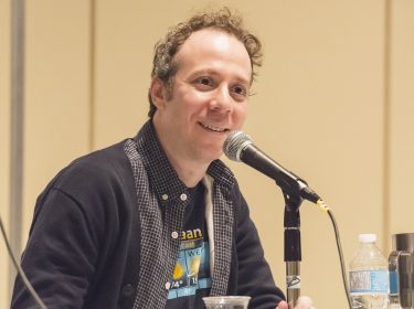 Auf dem Bild in dem Artikel zu dem Big Bang Theory-Spin-off „Stuart Fails to Save the Universe“ ist Kevin Sussman zu sehen, der Stuart in der Serie spielt. Er sitzt lächelnd bei einem Panel hinter einem Mikrofon, trägt ein dunkelblaues Hemd und eine Wettergrafik auf dem T-Shirt. Die Atmosphäre ist locker und freundlich.