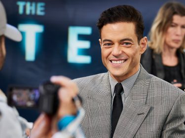 Auf dem Bild zur Premiere von The Amateur ist Schauspieler Rami Malek zu sehen. Er trägt ein elegantes, kariertes Sakko mit Krawatte und lächelt in die Kamera, während im Hintergrund Medienvertreter mit Kameras zu sehen sind. Das Bild gehört zu einer Liste der besten Filme von Rami Malek.