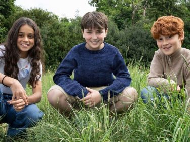 Auf dem Bild zur neuen Harry Potter-Serie sind drei Kinder in einer Wiesenlandschaft zu sehen. In der Mitte sitzt ein Junge mit dunkelblauem Pullover, links von ihm kniet ein Mädchen mit langen dunklen Haaren und weißem T-Shirt, rechts sitzt ein rothaariger Junge in einem beigefarbenen Pullover.