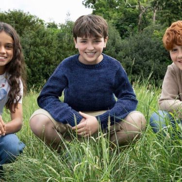Auf dem Bild zur neuen Harry Potter-Serie sind drei Kinder in einer Wiesenlandschaft zu sehen. In der Mitte sitzt ein Junge mit dunkelblauem Pullover, links von ihm kniet ein Mädchen mit langen dunklen Haaren und weißem T-Shirt, rechts sitzt ein rothaariger Junge in einem beigefarbenen Pullover.