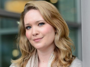 Auf dem Bild zur ACOTAR-Verfilmung ist die US-amerikanische Fantasy-Autorin Sarah J. Maas zu sehen, die „Das Reich der Sieben Höfe“ geschrieben hat. Sie blickt freundlich in die Kamera, trägt ihre blonden Haare offen und ist vor einem unscharfen Hintergrund mit Glasfassade zu sehen. Das Bild vermittelt eine ruhige, optimistische Stimmung und hebt die Autorin der Romanvorlage zur ACOTAR-Verfilmung hervor.