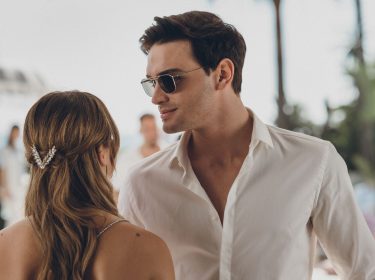 Diese Szene aus Culpa Nuestra zeigt Gabriel Guevara als Nick im weißen Hemd mit Sonnenbrille, wie er einer Frau – von hinten zu sehen, mit eleganter Haarspange – nahekommt. Die Atmosphäre wirkt sommerlich und leicht angespannt, mit Palmen und Meer im Hintergrund, was auf eine Hochzeit oder Feier am Strand hindeutet.