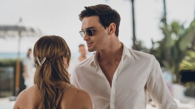 Diese Szene aus Culpa Nuestra zeigt Gabriel Guevara als Nick im weißen Hemd mit Sonnenbrille, wie er einer Frau – von hinten zu sehen, mit eleganter Haarspange – nahekommt. Die Atmosphäre wirkt sommerlich und leicht angespannt, mit Palmen und Meer im Hintergrund, was auf eine Hochzeit oder Feier am Strand hindeutet.