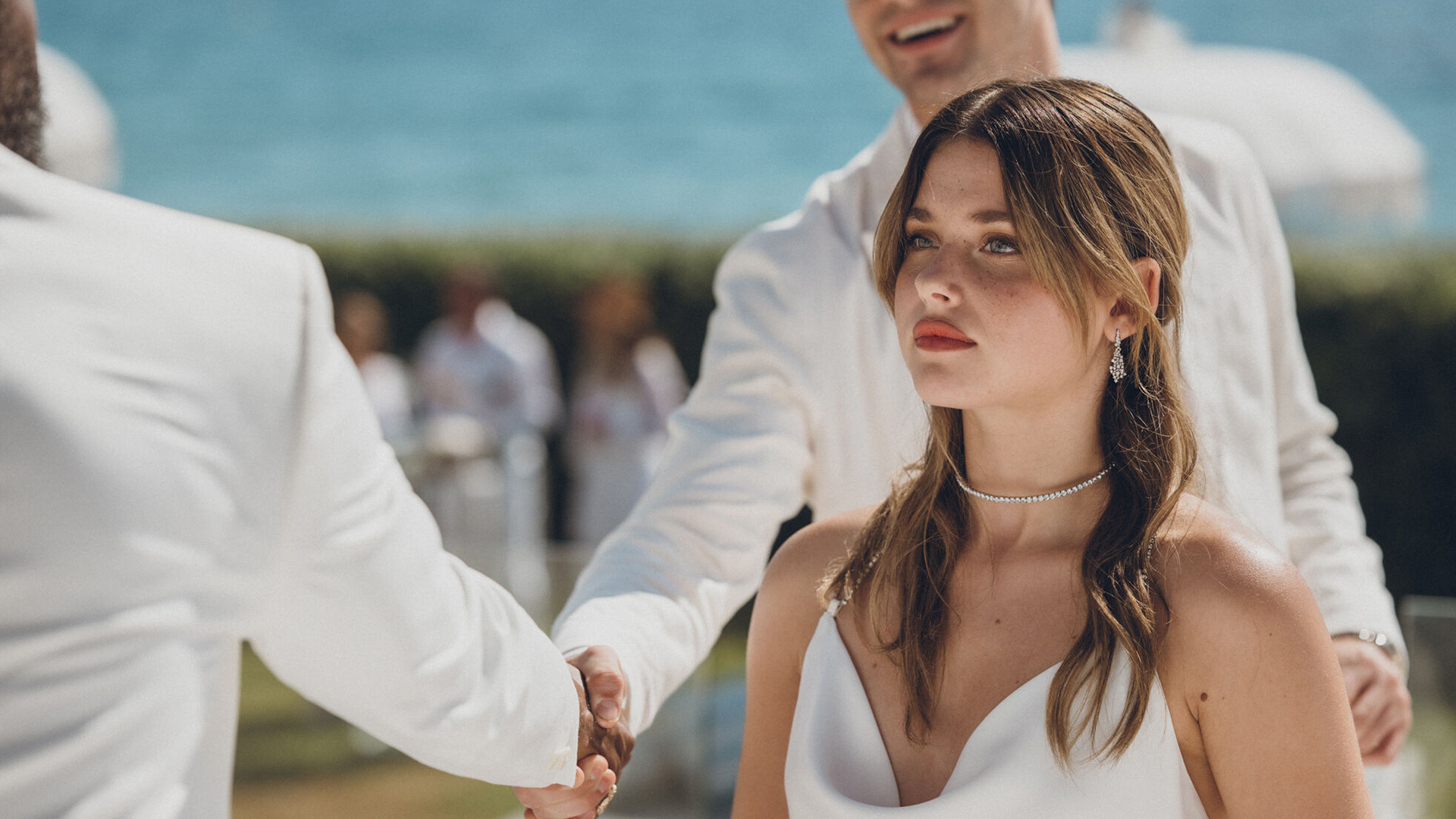 In dieser Szene aus Culpa Nuestra ist Nicole Wallace als Noah bei einer Hochzeit zu sehen. Sie trägt ein elegantes weißes Kleid, dezente Diamantohrringe und eine funkelnde Halskette. Ihr Ausdruck wirkt ernst und nachdenklich, während zwei Männer im Hintergrund in Weiß gekleidet Hände schütteln. Die Kulisse zeigt das Meer im Hintergrund und verleiht der Szene eine feierliche Stimmung.