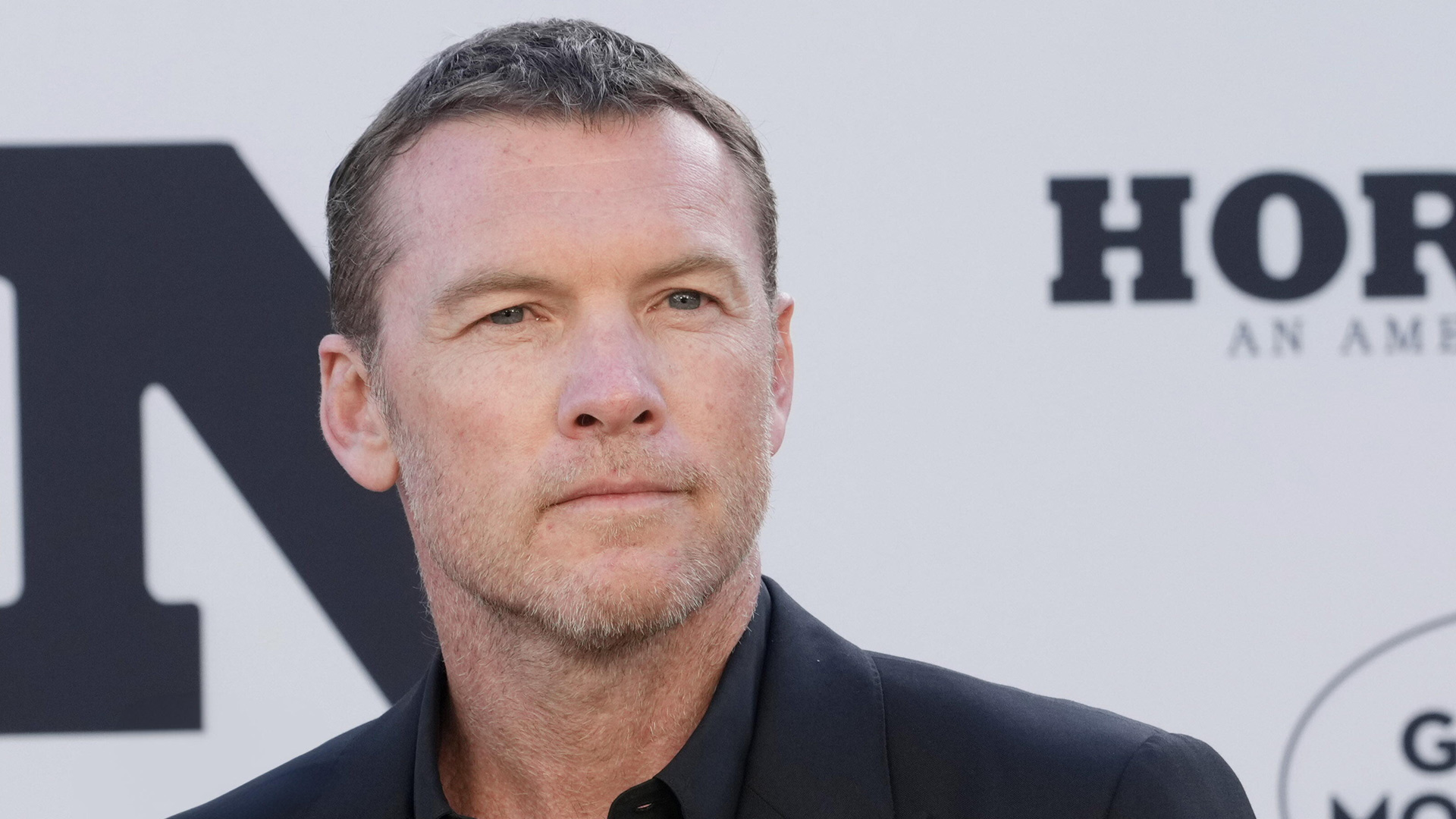 Auf dem Bild zur Nur für Dein Leben Netflix-Serie ist Sam Worthington in einem eleganten schwarzen Anzug vor einer neutralen Hintergrundwand mit großem Schriftzug zu sehen. Er blickt ruhig und ernst zur Seite, sein Gesichtsausdruck wirkt konzentriert und nachdenklich.