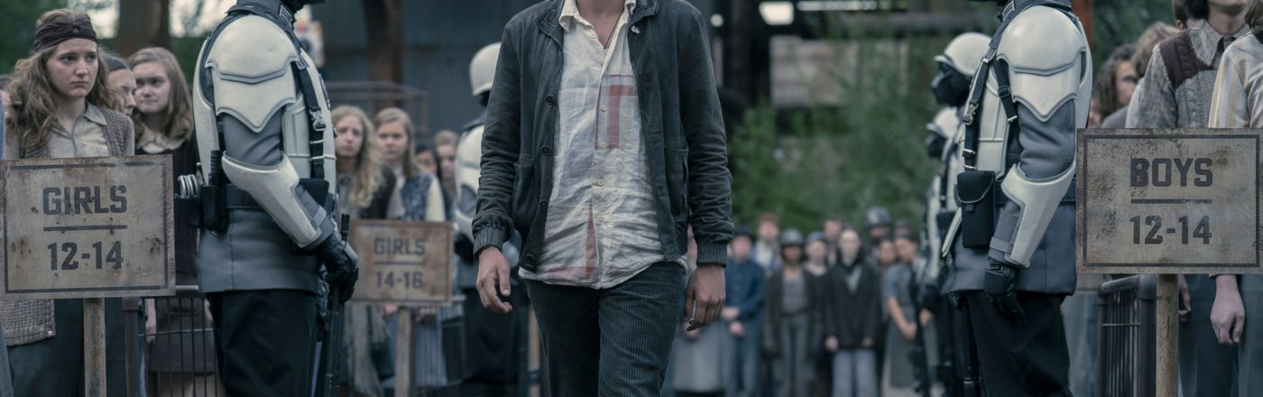 Im Bild zu Tribute von Panem 6 schreitet Joseph Zada als junger Haymitch Abernathy entschlossen durch eine Kontrollzone der Hungerspiele. Links und rechts stehen Peacekeeper in weißen Rüstungen, während Jugendliche nach Altersgruppen getrennt in Reihen stehen. Die Szene wirkt angespannt und symbolisiert den Moment der Auslese mit düsterer, unterdrückender Stimmung.