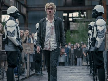 Im Bild zu Tribute von Panem 6 schreitet Joseph Zada als junger Haymitch Abernathy entschlossen durch eine Kontrollzone der Hungerspiele. Links und rechts stehen Peacekeeper in weißen Rüstungen, während Jugendliche nach Altersgruppen getrennt in Reihen stehen. Die Szene wirkt angespannt und symbolisiert den Moment der Auslese mit düsterer, unterdrückender Stimmung.