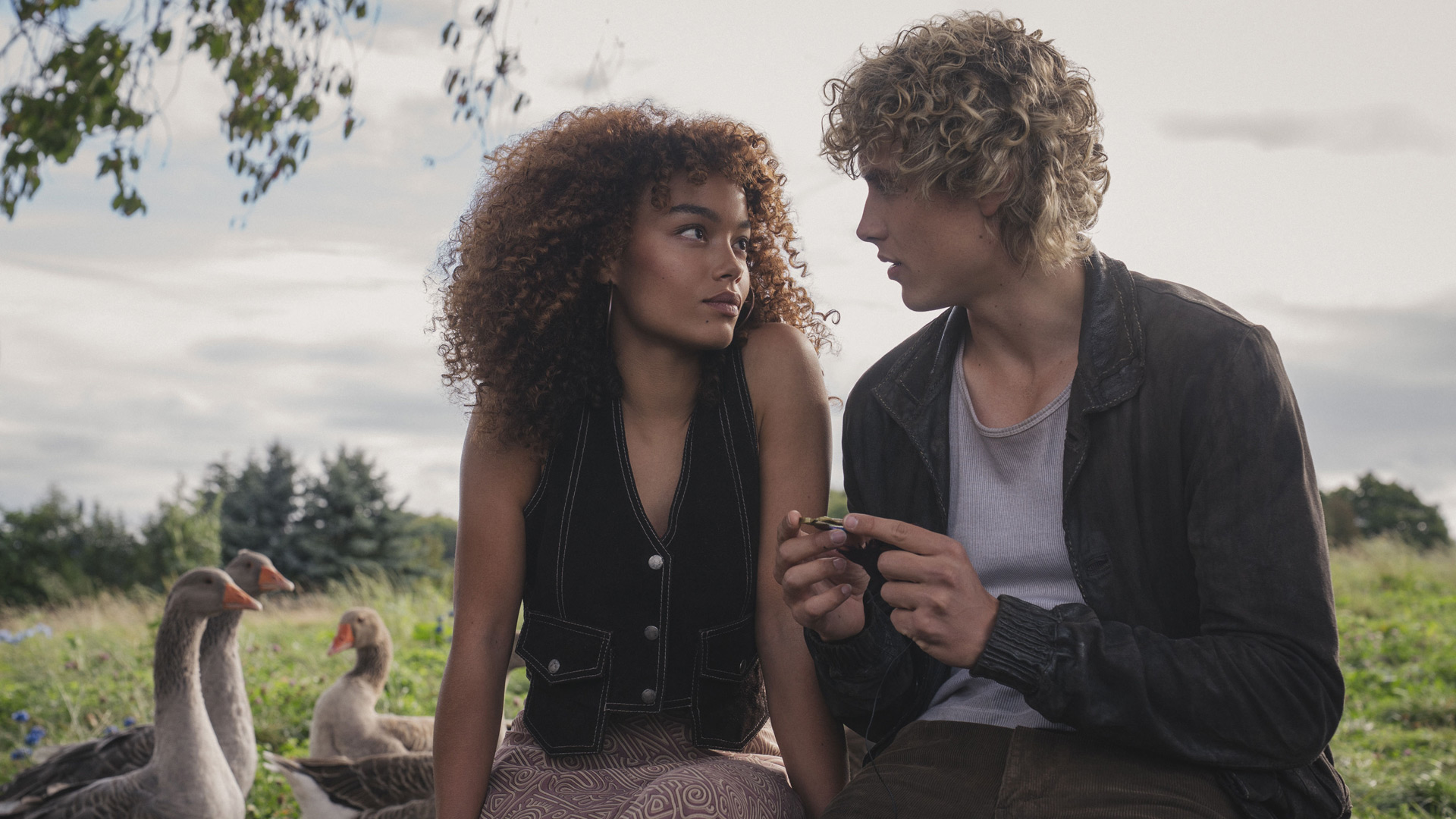 Auf dem Bild zu Tribute von Panem 6 sind Whitney Peak als Lenore Dove und Joseph Zada als junger Haymitch Abernathy in einer ruhigen, nachdenklichen Szene zu sehen. Die beiden sitzen auf einer Wiese, umgeben von Gänsen, und blicken sich intensiv an. Die ländliche Umgebung wirkt friedlich und steht im Kontrast zur angespannten Atmosphäre zwischen den Charakteren.