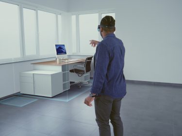 Auf dem symbolischen Bild zum Thema „Was ist Mixed Reality“ steht ein Mann mit Mixed-Reality-Brille in einem leeren Raum und interagiert mit einem digital eingeblendeten Schreibtisch samt Monitor, Stuhl und Kaffeetasse. Die Szene zeigt beispielhaft, wie virtuelle Objekte nahtlos in die reale Umgebung projiziert werden und veranschaulicht das Potenzial von Mixed Reality im Arbeitsalltag.