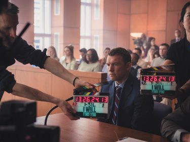 Auf diesem Bild zur Serie Nur für Dein Leben ist Sam Worthington als David Burroughs in einer Gerichtsszene zu sehen. Er sitzt ernst und fokussiert am Tisch, umgeben von Filmcrew-Mitgliedern mit Klappe, was auf eine laufende Produktion hinweist. Die Atmosphäre ist angespannt, mit Statisten im Hintergrund, die ein authentisches Gerichtssetting verstärken.