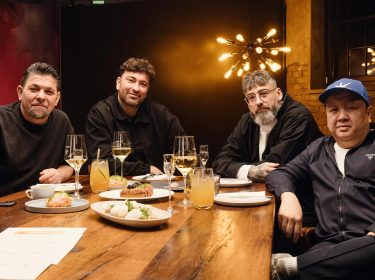 In dieser Szene aus Kitchen Impossible Staffel 10 sitzen Tim Mälzer, Marteria, Sido und The Duc Ngo gemeinsam an einem festlich gedeckten Tisch.