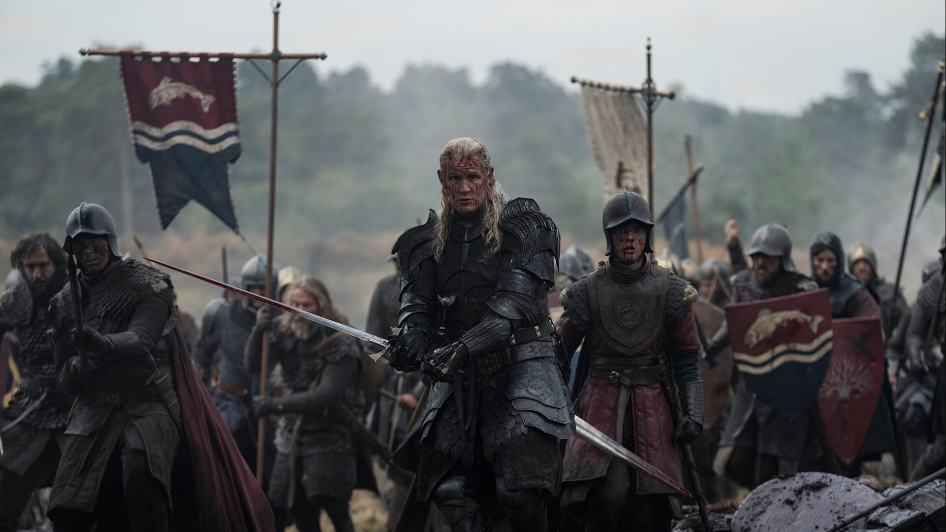 Auf dem Bild zu House of the Dragon Staffel 3 ist Matt Smith als Daemon Targaryen in schwarzer, kampferprobter Rüstung mit gezogenem Schwert zu sehen. Er steht entschlossen an vorderster Front eines mittelalterlichen Schlachtfelds, umgeben von schwer gerüsteten Soldaten mit blutverschmierten Gesichtern. Die Szene wirkt düster, angespannt und voller Dramatik.