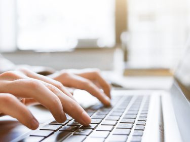 Auf dem Bild in dem Artikel zum Erstellen von ChatGPT-Bildern sind zwei Hände zu sehen, die auf der Tastatur eines Laptops tippen. Die Szene ist in weichem Tageslicht gehalten, das durch ein Fenster im Hintergrund fällt.