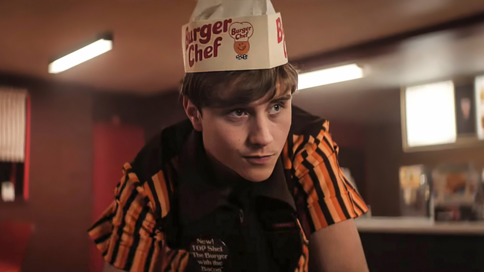 Dieses Symbolbild zu Die Tribute von Panem 6 zeigt Joseph Zada, der im neuen Film die Rolle des Haymitch Abernathy übernimmt. Er trägt eine Burger-Chef-Uniform aus dem Film The Speedway Murders, inklusive gestreiftem Hemd und Papierhut. Die Szene ist in einem Fast-Food-Restaurant angesiedelt und wirkt durch das Licht nostalgisch und retro.