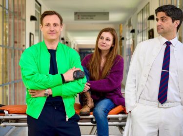 Auf dem Bild zur Serie Dr. Nice Staffel 4 stehen drei Hauptfiguren in einem Krankenhausflur. Links steht Dr. Nice, gespielt von Patrick Kalupa, in einer auffälligen grünen Jacke und lächelt selbstbewusst. In der Mitte sitzt Charlie Winkler, dargestellt von Josefine Preuß, locker auf einer Trage und blickt freundlich. Rechts lehnt Dr. Florian Schmidtke, gespielt von Maximilian Grill, in einem weißen Arztkittel mit verschränkten Armen und skeptischem Blick.