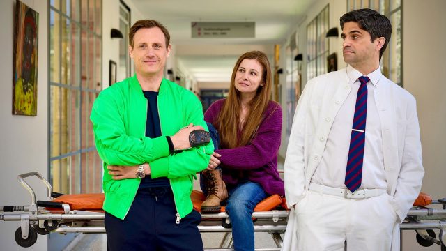 Auf dem Bild zur Serie Dr. Nice Staffel 4 stehen drei Hauptfiguren in einem Krankenhausflur. Links steht Dr. Nice, gespielt von Patrick Kalupa, in einer auffälligen grünen Jacke und lächelt selbstbewusst. In der Mitte sitzt Charlie Winkler, dargestellt von Josefine Preuß, locker auf einer Trage und blickt freundlich. Rechts lehnt Dr. Florian Schmidtke, gespielt von Maximilian Grill, in einem weißen Arztkittel mit verschränkten Armen und skeptischem Blick.