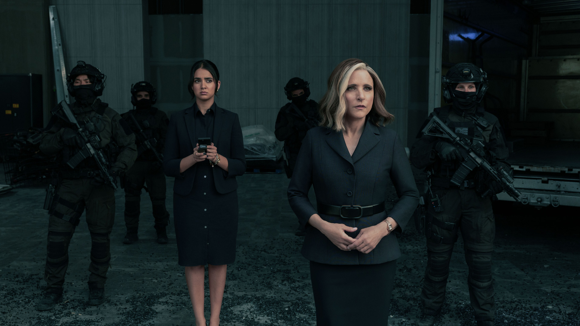 Im Bild zur Thunderbolts* Kritik steht Valentina Allegra de Fontaine, gespielt von Julia Louis-Dreyfus, im Vordergrund mit ernster Miene. Rechts und links von ihr sind schwer bewaffnete Sicherheitskräfte zu sehen. Neben ihr steht eine junge Frau, dargestellt von Geraldine Viswanathan, die auf ein Gerät schaut. Die Szene wirkt angespannt und professionell, mit düsterem Licht und kühler Farbgebung.