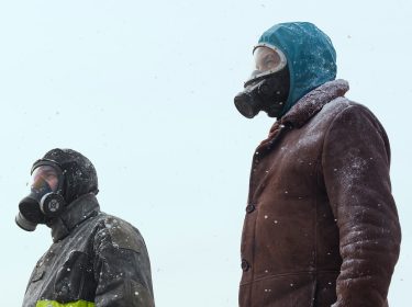 In dieser Szene aus Eternauta Staffel 1 stehen zwei Figuren in einer verschneiten Außenlandschaft mit Gasmaske und Winterkleidung. Links ist Favalli zu sehen, gespielt von César Troncoso, in dunkler Jacke mit reflektierendem Streifen. Rechts steht Juan, dargestellt von Ricardo Darín, in brauner Felljacke und Kapuze. Beide blicken ernst in die Ferne – die Stimmung ist angespannt und bedrohlich.