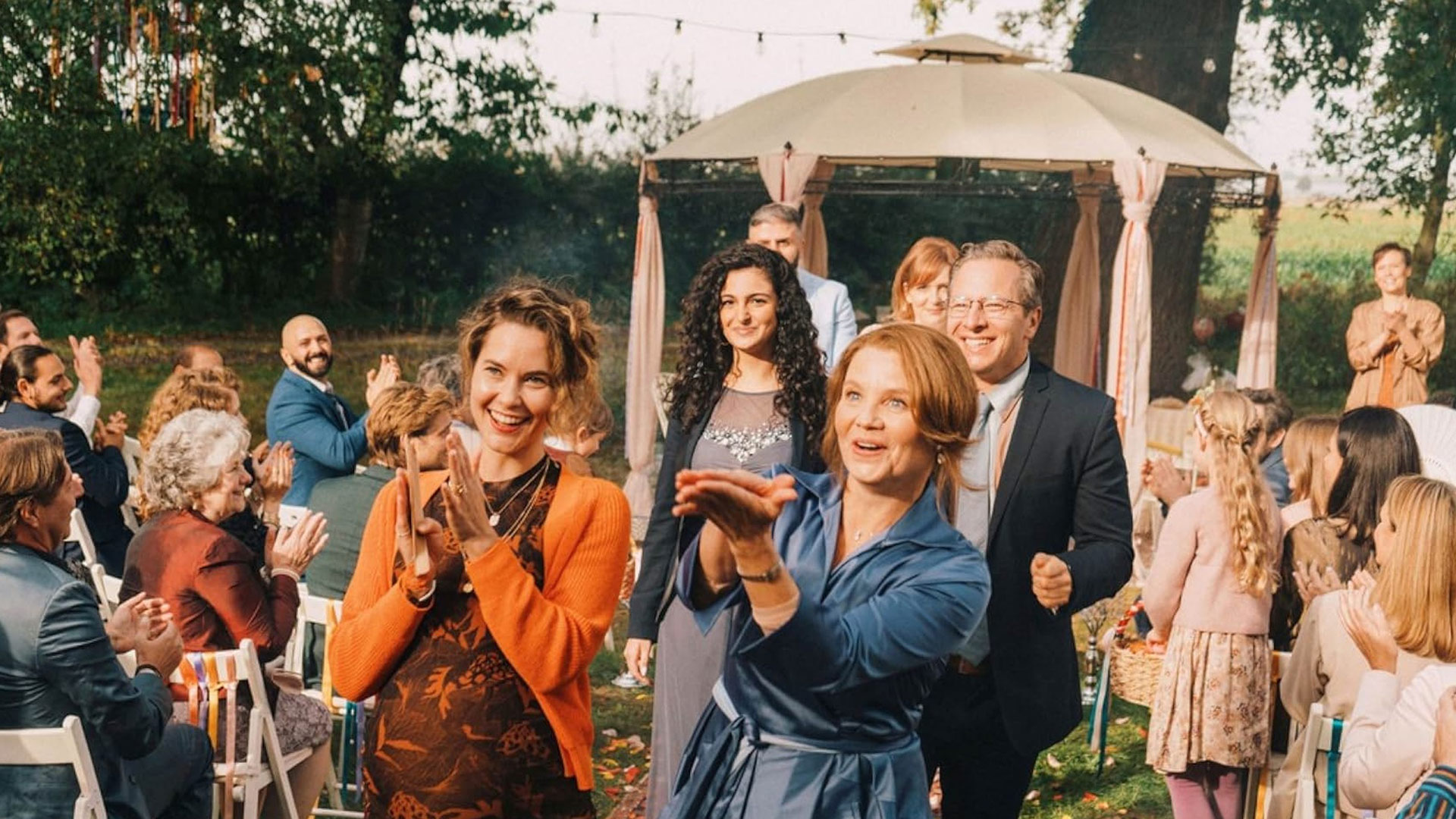 Das Bild zu den GigaTV-Highlights im Mai stammt aus dem Film „Feste & Freunde“. In einer fröhlichen Szene feiern zahlreiche Menschen gemeinsam eine Gartenhochzeit. Im Vordergrund applaudieren zwei Frauen begeistert, umgeben von lachenden Gästen, bunten Kleidern und festlicher Dekoration. Die Atmosphäre ist herzlich, familiär und voller Lebensfreude.