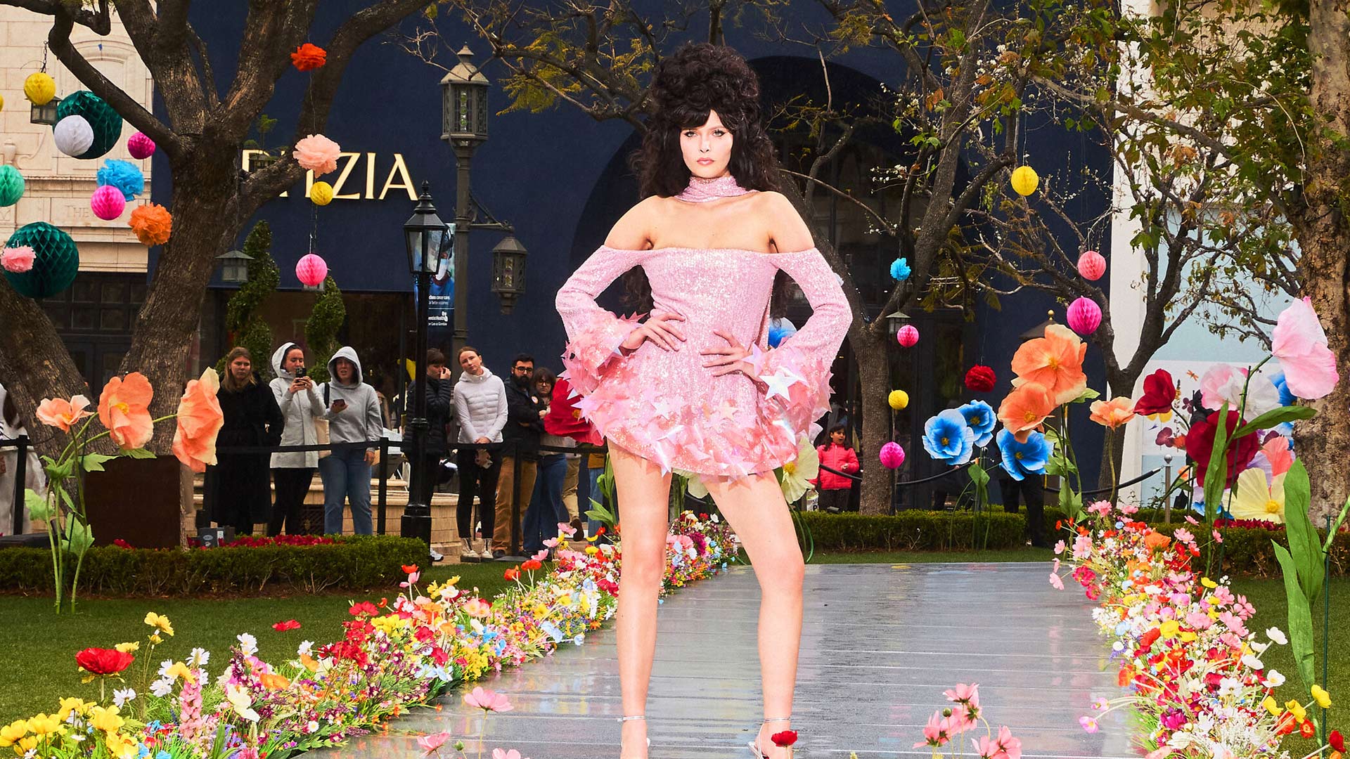 Auf dem Bild zur GNTM 2025 ist Kandidatin Magdalena beim großen Walk vor Live-Publikum zu sehen. Sie trägt ein funkelndes, rosafarbenes Minikleid mit schulterfreiem Schnitt, designt von Stardesigner Christian Cowan. Die mit Sternen verzierte Szenerie ist bunt geschmückt, das Publikum im Hintergrund schaut gespannt zu. Die Stimmung wirkt festlich und selbstbewusst.