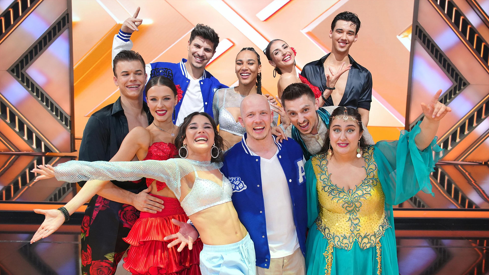 Auf dem Bild zu Lets Dance 2025 sind zehn Tänzer:inne fröhlich und energiegeladen vor einem leuchtenden Bühnenhintergrund zu sehen. Alle tragen auffällige Kostüme – von glitzernden Tops über bunte Tanzkleider bis zu sportlichen Collegejacken. Die Stimmung wirkt ausgelassen und sie lachen direkt in die Kamera.